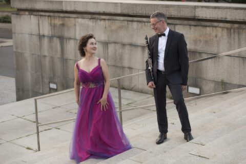 Musiker und Sängerin bei einem Konzert im Freien, elegante Kleidung, Stadtarchitektur im Hintergrund.